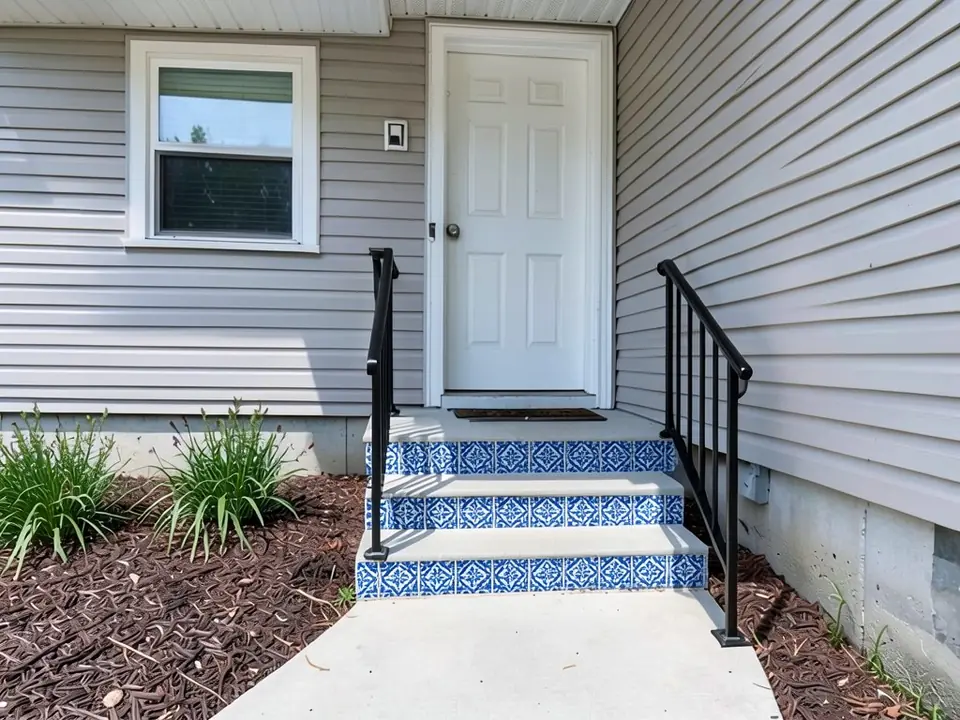The same steps with blue and white patterned tile on the risers, plain concrete treads and new black metal handrails on both sides; door, siding, window and plants unchanged