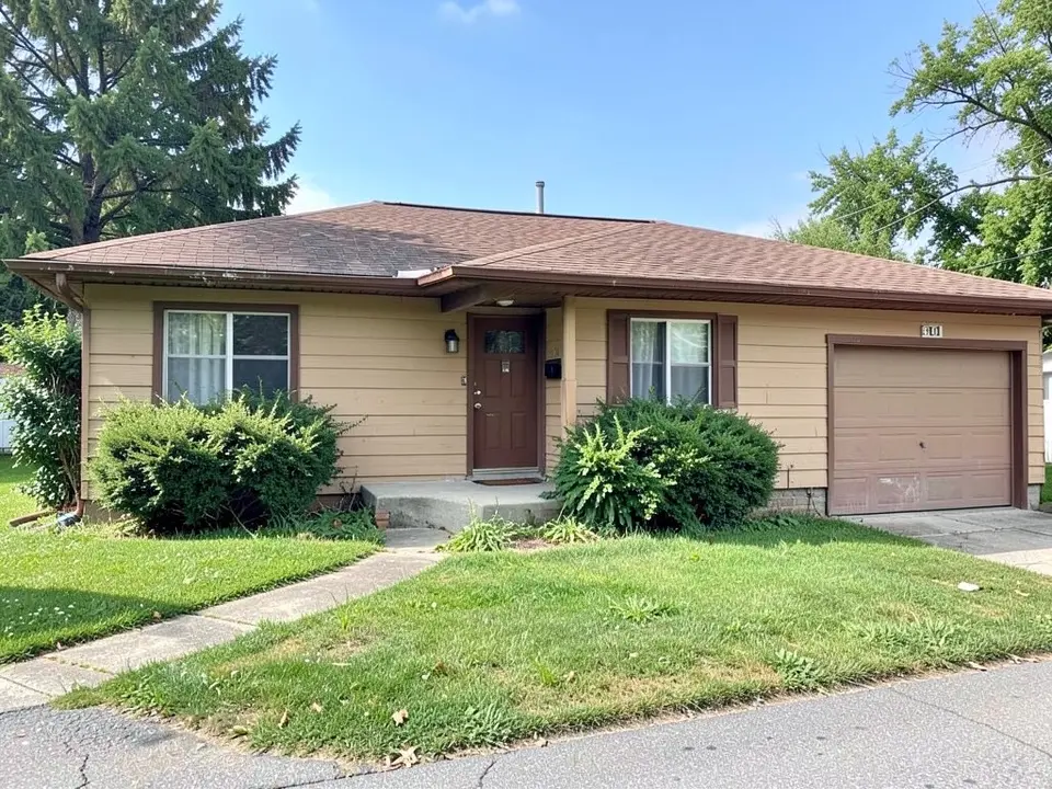 Dated single-story ranch house with tan siding, brown trim, a brown front door and overgrown shrubs