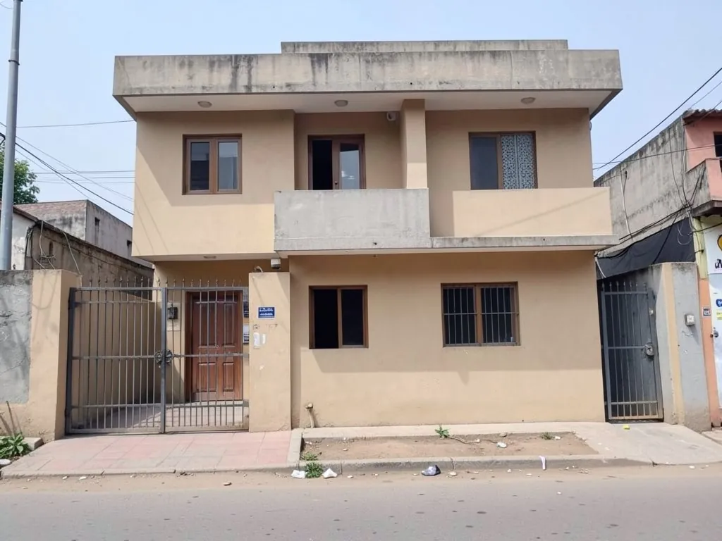 AI-drawn generic stand-in of a plain two-story house front with a flat roof, beige plaster walls, a concrete balcony and an iron gate on a narrow street