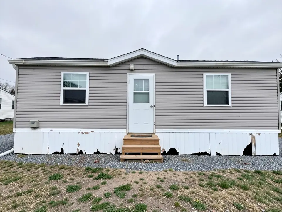 Single-wide manufactured home with grey vinyl siding, a white front door and wooden steps, and old white vinyl skirting with torn and missing panels showing dark gaps underneath