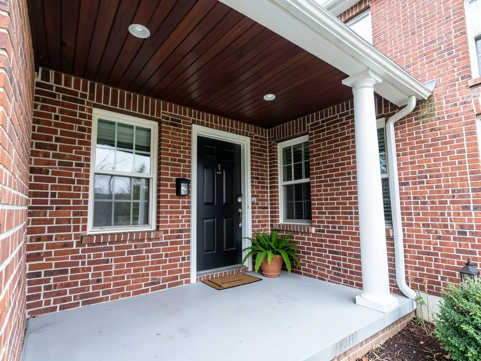 AI-drawn generic stand-in of a red brick house front porch seen from below, with a dark brown stained wood ceiling, a white column, a gray floor, a black front door and a potted fern