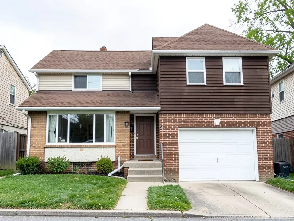 Split-level style house with a raised brick lower floor and basement window on the left, garage at street level on the right, brown and beige siding above and a white one-car garage door